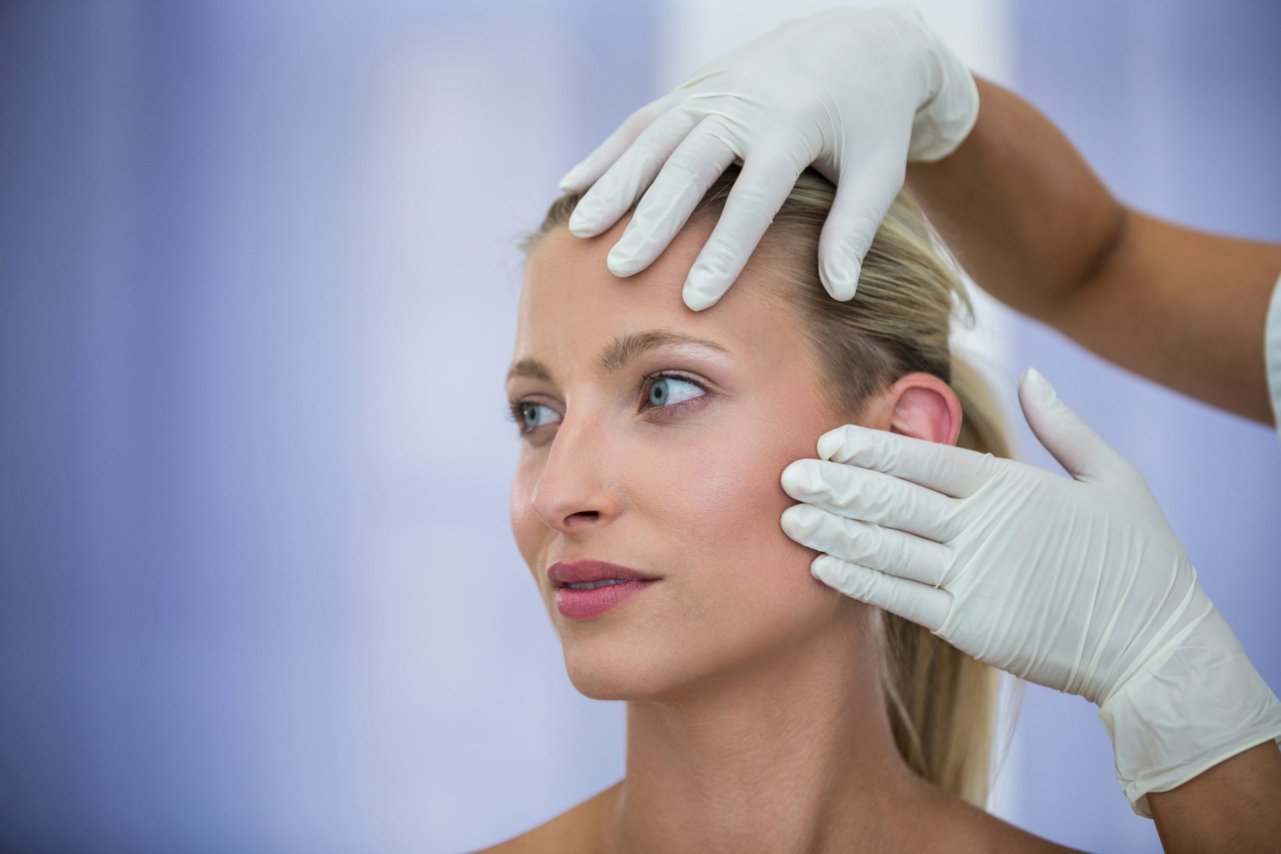 A close-up of a blonde woman looking off-camera while a practitioner wearing white medical gloves gently assesses the skin elasticity on her forehead and cheek.